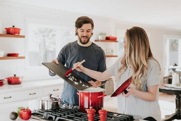Couple souriant cuisinant ensemble, versant des oignons dans une marmite rouge sur un piano de cuisson moderne.