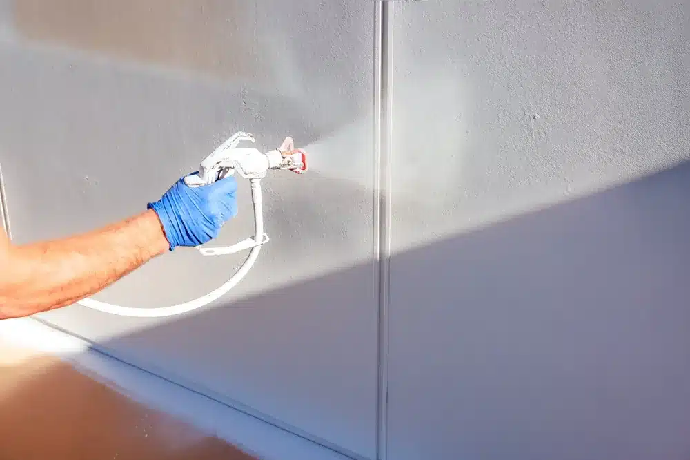 Peintre utilisant un pistolet pulvérisateur airless pour appliquer de la peinture blanche sur un mur intérieur.