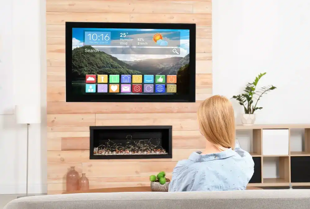 Femme devant une smart TV affichant 10:16 et 25°C, fixée sur un mur en bois au-dessus d'une cheminée moderne.