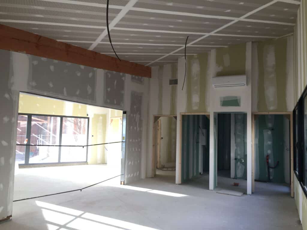 Travaux de plâtrerie et aménagement intérieur en cours Chantier intérieur : murs en plaques de plâtre, clim installée, poutre apparente et grandes fenêtres lumineuses.